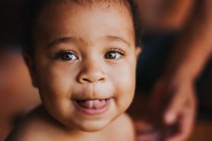 Um bebê sorri enquanto olha para a câmera, com atenção. Ao fundo, está os braços de um adulto em fundo desfocado. 